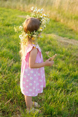 Fototapeta premium small child, happy cool blonde girl 4 years old in pink dress stands in green meadow, kid's head is decorated with cute wreath wildflowers, daisies, grass doll in hands, happy childhood