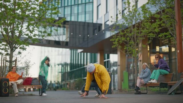 Static Shot of Young Stylish B-boy Breakdancing On Street Among Buildings In Urban Area. Fashionable Group Of Friends Chilling On Background, Supporting Breakdance Performer Practising Choreography.