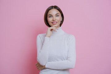 Beautiful woman, pleased expression, chin touch, tender smile, straight dark hair, white turtleneck, pink background. Femininity, beauty, skin care