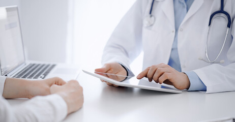 Doctor and patient sitting opposite each other at the desk in clinic. The focus is on female physician's hands using tablet computer, close up. Medicine concept