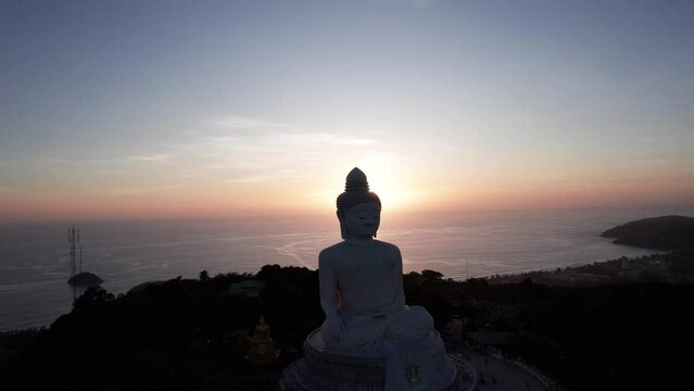 Big Buddha at sunset view from a drone. Phuket. People climb the steps to the statue. The green hills of the island are all around. In the distance, the bright sun goes over the sea. View from above