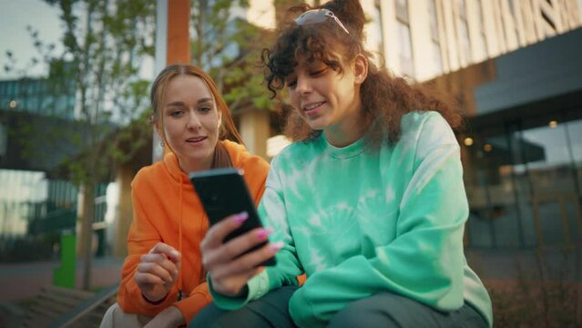 Two Young And Stylish Female Students Talking On City Street And Using Smartphone To Scroll Social Media Posts. Two Trendy Women Discussing University Assignments Latest News And Fashion Downtown.