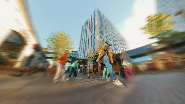 Trendy Zoom Blur Edit of Young B-boy Breakdancing On City Street Among Modern Buildings In Urban Area. Group Of Friends Cheering On Background, Supporting Breakdancer Practising Choreography.