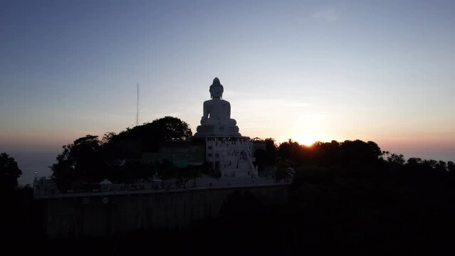 Big Buddha at sunset view from a drone. Phuket. People climb the steps to the statue. The green hills of the island are all around. In the distance, the bright sun goes over the sea. View from above