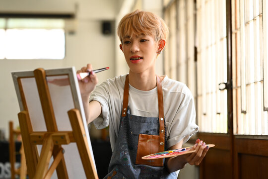 Inspired asian male student holding palette painting picture on canvas in art workshop. Art, education and leisure activity concept