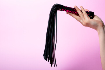 female hand holds a black leather whip with a pink handle on a pink background. Place for text. Sex shop concept. Copy space