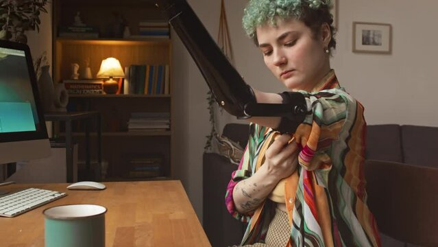 Caucasian girl in her early 20s with short curly hair putting on arm prosthesis while sitting at desk in front of computer