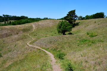 A view of a winding path leading through hills and slopes seen in the middle of a field, meadow, or pastureland, with some forests, moors, and wooden fences visible in the background in Poland