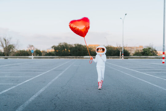 Girl wearing astronaut dress walking in parking lot holding red heart balloon