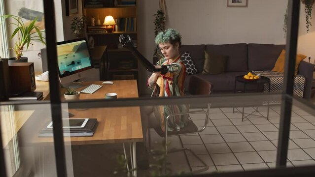 Medium shot of Caucasian girl with disability sitting by desk in front of window in cozy apartment attaching her arm prosthesis