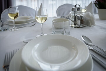 Biały stół ozdobiony białymi talerzami, serwetkami i kieliszkami do wina, ze sztućcami,...