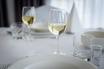 Biały stół ozdobiony białymi talerzami, serwetkami i kieliszkami do wina, ze sztućcami, ustawiony na imprezy - obrazy, fototapety, plakaty