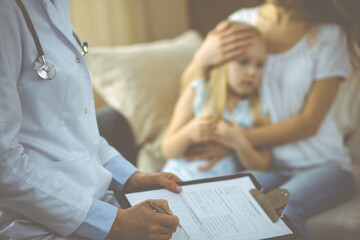 Doctor and patient. Pediatrician using clipboard while examining little girl with her mother at home. Sick and unhappy child at medical exam. Medicine concept