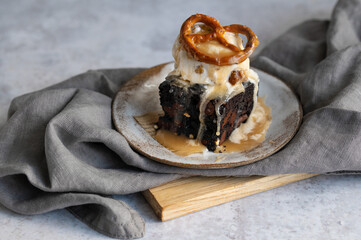Dark chocolate brownies with 
scoop of ice cream and salted caramel sauce and pretzels on a grey stone background. Dessert from American cuisine.