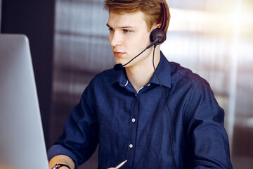 Young blond businessman using headset and computer in a darkened office, glare of light on the background. Startup business means working hard