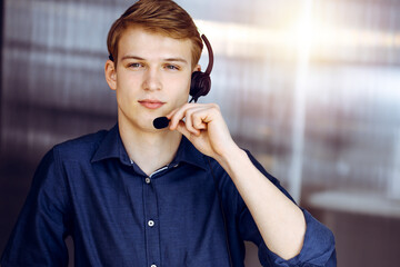 Young blond businessman using headset and computer in a darkened office, glare of light on the background. Startup business means working hard