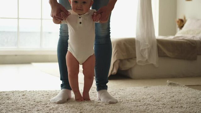 Baby development. Little infant girl making first steps at home. Mother holding baby hands helping toddler child to take first steps walking in living room. Happy family at home. Mom teaching baby