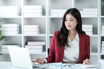 Beautiful asian businesswoman working on laptop and calculating with financial calculator from statistics data graphs, charts. Successful business results in modern office wear red shirt.