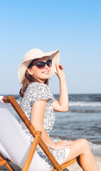 Happy brunette woman wearing sunglasses while relaxing on a wooden deck chair at the ocean beach
