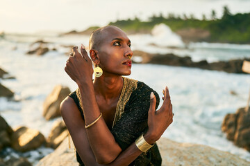 Non-binary black person in luxury dress sits on rocks in ocean. Trans ethnic fashion model wearing...