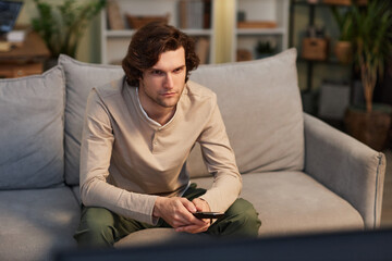 Portrait of young man watching TV at home sitting on sofa in cozy green interior, copy space