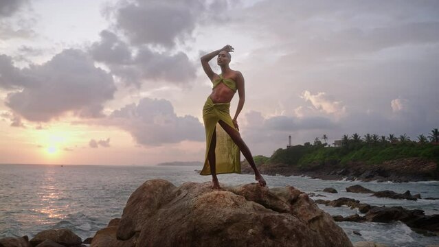 LGBTQIA black person in open dress, jewelry stands on rock on stunning ocean coastline sunset. Gender fluid ethnic divine model in seductive outfit fluttering in the wind against dramatic sky.