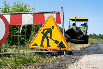 Road work ahead warning sign, hanging on a fence of construction site of new road. Paver finisher...
