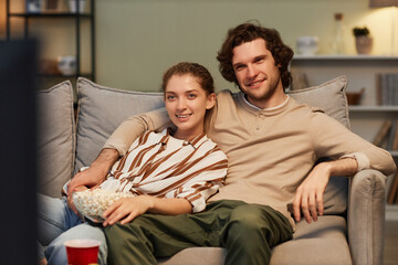 Portrait of smiling couple watching movies at home and eating popcorn sitting on couch together