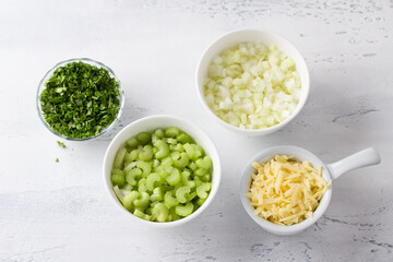 Bowls with chopped stalk celery, onion, parsley and grated cheese on a light blue background, top view. Cooking a delicious vegetarian casserole, soup or other dish