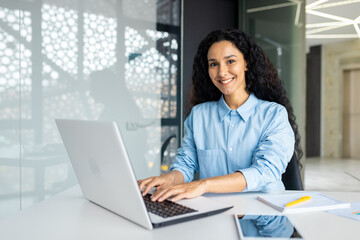 Fototapeta na wymiar Young beautiful Indian female programmer working inside the office on a laptop, woman smiling and looking at the camera, portrait of a satisfied and confident businesswoman at the workplace.