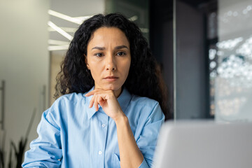 Fototapeta na wymiar Serious thinking woman working inside office at workplace with laptop, confident hispanic woman looking at technical task, brainstorming solution, business woman with curly hair looking at laptop