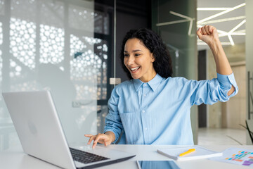 Fototapeta na wymiar Successful joyful businesswoman satisfied with results of achievement at workplace, Latin American received online feedback, holds hand up celebrating victory success, triumph gesture.