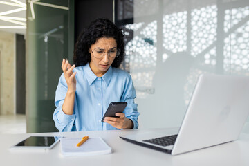 Shocked and upset business woman working inside office at workplace, depressed hispanic woman reading bad news using app on smartphone, holding phone.