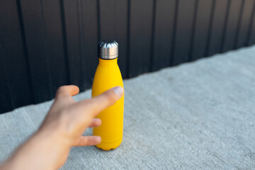 Close-up of male hand want to take a steel thermo bottle of yellow color from the concrete floor.