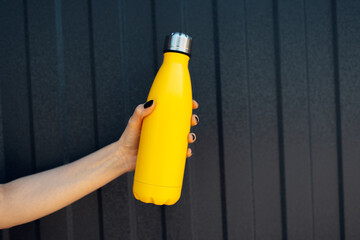Female hand holding a steel thermo water bottle of yellow color on metal background with lines of black color.
