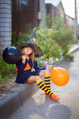 Fototapeta na wymiar A little girl in a witch costume and with balloons for Halloween walks down the street and rejoices in the holiday