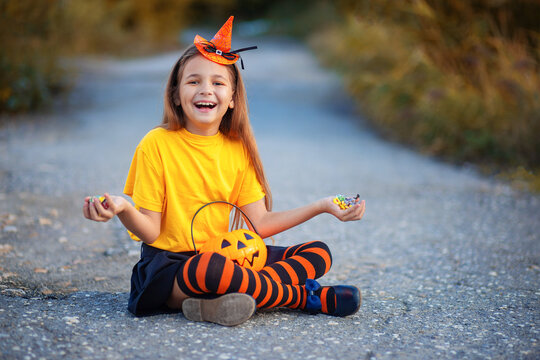 A girl in a Halloween costume sits on the ground and enjoys candy from her basket. The child rejoices in sweets for Halloween