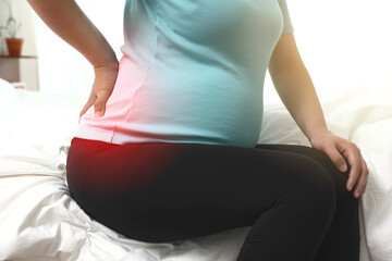 임신한 여성이 자신의 아픈 허리를 만지며 침대에 앉아있다(붉은색 강조)