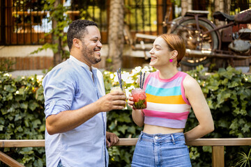 Fototapeta premium A multi-ethnic couple happily drinks mojitos on the terrace of a bar. African man and Caucasian woman look at each other while drinking alcoholic cocktails. Interracial love.