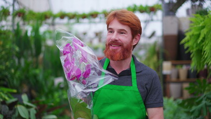 Happy young man employee of plant store carrying flower. A male redhead caucasian person wearing green apron walks through business store aisle