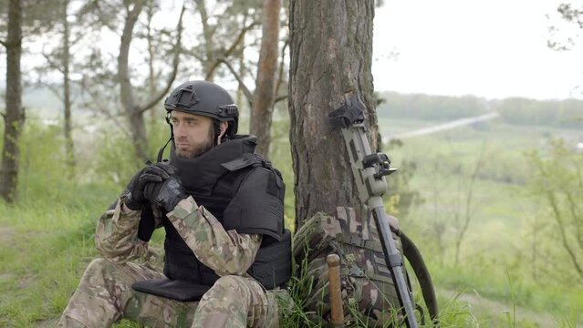 Mine clearance. Soldier with mine detector and backpack sits exhausted by a tree after heavy demining. Demining process