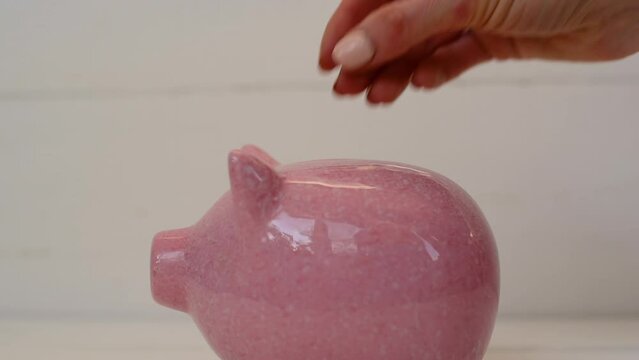 Pink piggy bank mumps on white background. Womans hand slowly puts coin into piggy bank. Cash savings in future. Close-up