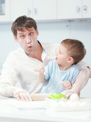 Father and son are preparing dinner in kitchen. Child has soiled his father with flour and is laughing. Fun photo of parent cooking with their little son.