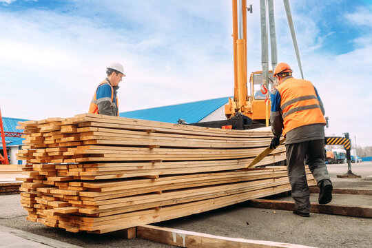 Two workers in helmets and construction vests unload wooden planks. Slingers stack lumber outside in the open air. Loading and unloading works. Authentic workflow.