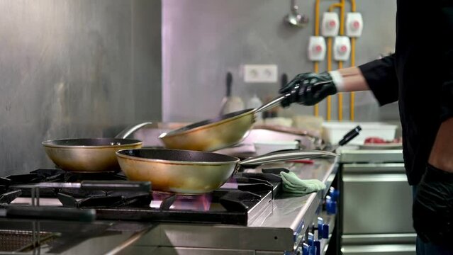 Real time side view of crop anonymous male cook in latex gloves flipping food in frying pans while preparing tasty dishes for clients in professional restaurant kitchen