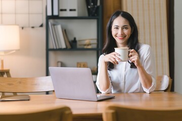 A millennial Asian businesswoman is sitting and working with laptop computer in her home kitchen, while also enjoying a relaxing cup of coffee.