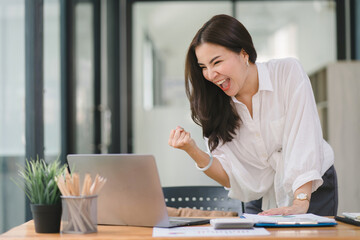 Portrait of a happy young businesswoman celebrating success with arms raised in front of a laptop,...
