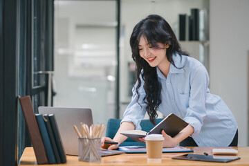 Smiling young Asian businesswoman holding a notebook, working in an office. Professional executive manager or saleswoman using corporate technology.