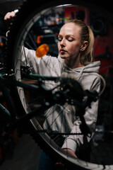 Fototapeta na wymiar Vertical front view of blonde cycling mechanic female repairing and fixing mountain bicycle standing on bike rack in repair workshop with dark interior. Concept of professional bicycle maintenance.