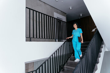 Brunette female doctor in blue uniform working at hospital, walking down stairs at modern clinic interior. Medicine, profession and healthcare concept.